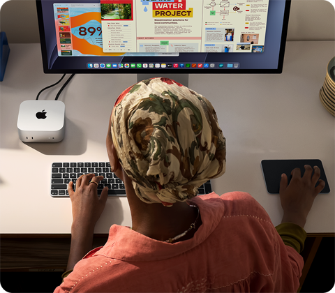 Човек пише на бюро с Mac mini, свързан със Studio Display, Magic Keyboard, Magic Trackpad, различни неща