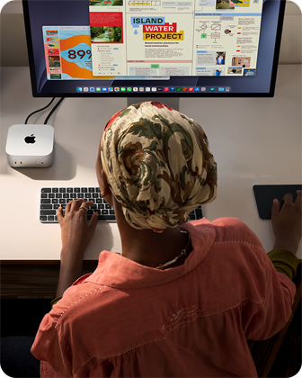Човек пише на бюро с Mac mini, свързан със Studio Display, Magic Keyboard, Magic Trackpad, различни неща
