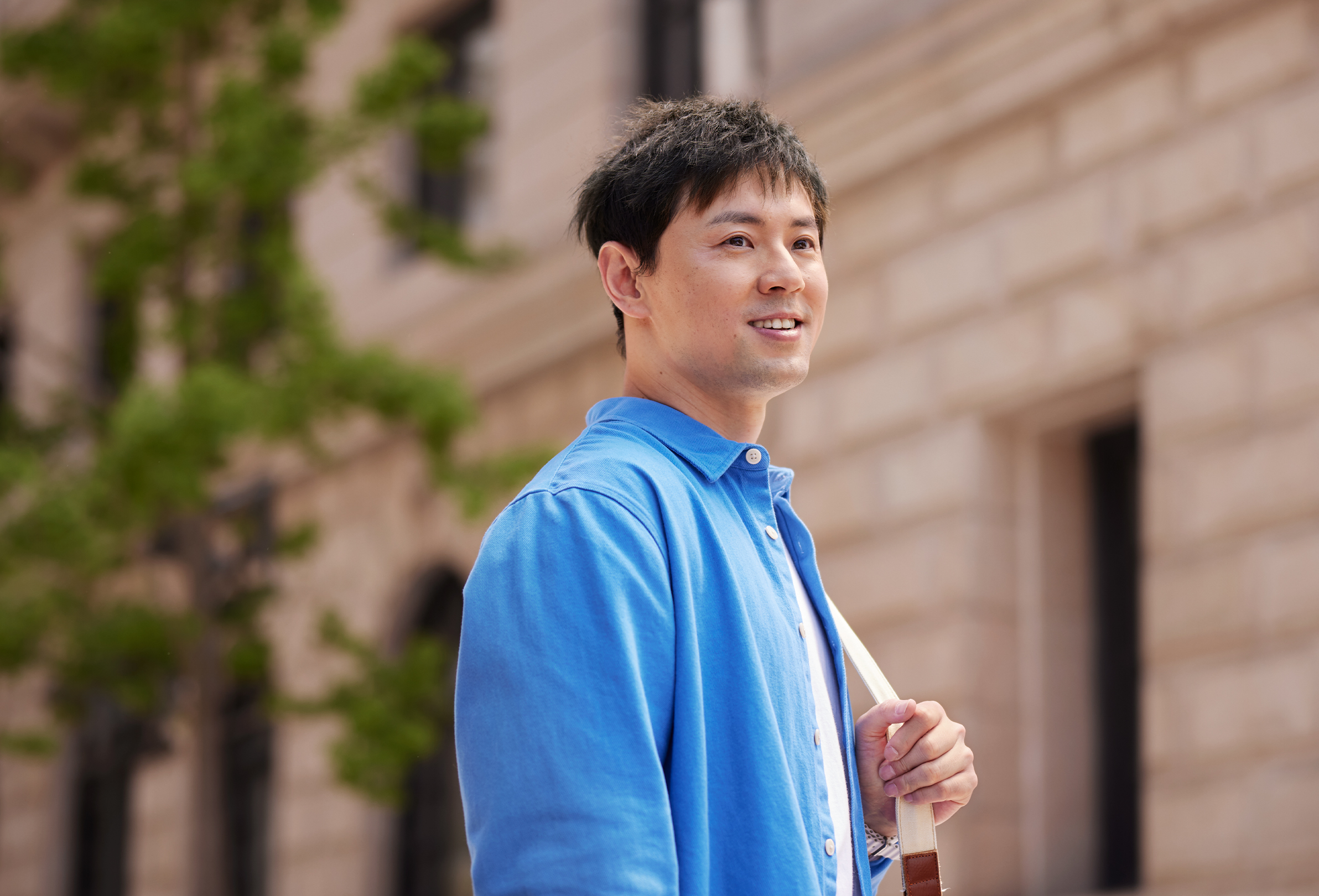 พนักงาน Apple ยืนยิ้มอยู่ด้านนอกอาคารสำนักงานก่อด้วยอิฐ มีกระเป๋าสะพายไหล่
