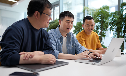 Ken กำลังพิมพ์ MacBook มีเพื่อนร่วมงานนั่งข้างๆ ด้านหลังมีหน้าต่างและต้นไม้เขียว