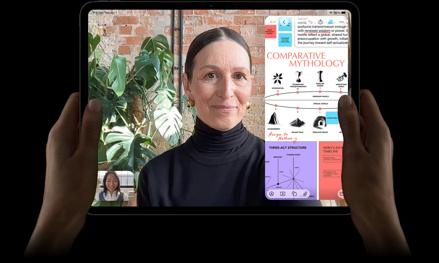 Vooraanzicht van iPad Pro in de kleur spacezwart, linker- en rechterhand houden het device in horizontale stand vast, afgeronde hoeken, zwarte rand rond het display, op het display wordt een FaceTime-gesprek live vertaald en in een educatieve app is een presentatie te zien over vergelijkende mythologie met tekst, diagrammen en handgeschreven notities