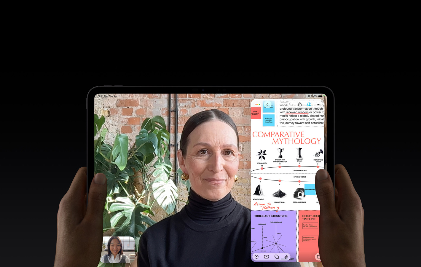 Vooraanzicht van iPad Pro in de kleur spacezwart, linker- en rechterhand houden het device in horizontale stand vast, afgeronde hoeken, zwarte rand rond het display, op het display wordt een FaceTime-gesprek live vertaald en in een educatieve app is een presentatie te zien over vergelijkende mythologie met tekst, diagrammen en handgeschreven notities