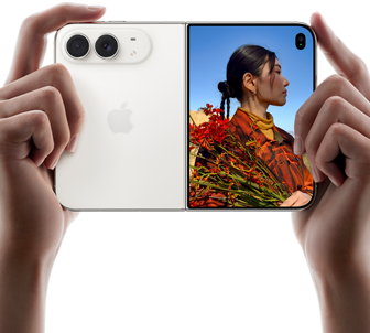 iPhone&nbsp;Duo, zvaigžņu baltā krāsā, tiek turēts divās rokās horizontālā novietojumā, atvērts, kreisajā panelī redzamas 48&nbsp;MP Dual&nbsp;Fusion aizmugurējās kameras, labajā panelī redzams, kā ārējā displejā tiek uzņemta cilvēka bilde, fotografējamais redz savu seju, kamēr tiek uzņemta bilde ar Duo&nbsp;Preview