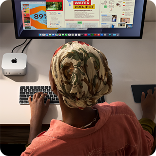 Niekto píše pri stole, na ktorom je Mac mini s pripojeným displejom Studio Display, klávesnica Magic Keyboard, trackpad Magic Trackpad a rôzne predmety