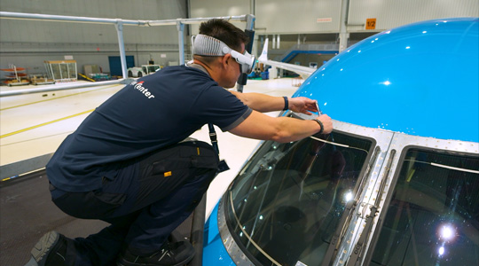 A technician in a plane hangar wears Apple Vision Pro while using a tool to adjust the plane’s windshield