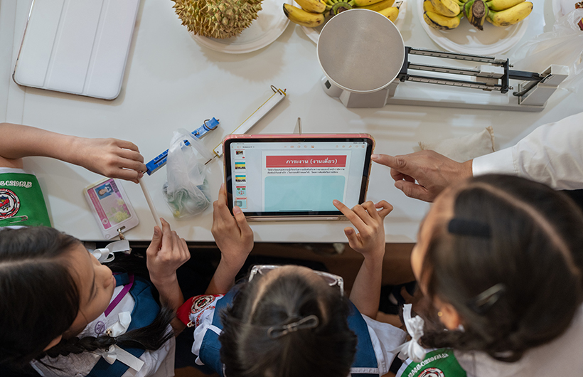 Três alunos do ensino básico reunidos em torno de um iPad para colaborar numa apresentação para um projeto de ciências.