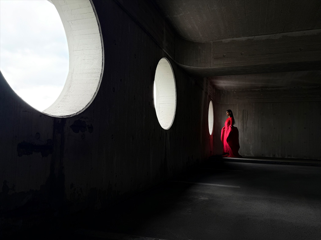 A woman wearing a long red dress stands in an empty room being photographed. The Photo app toggles through the focal lengths to demonstrate the capabilities of the pro camera system, showcasing: 28 mm